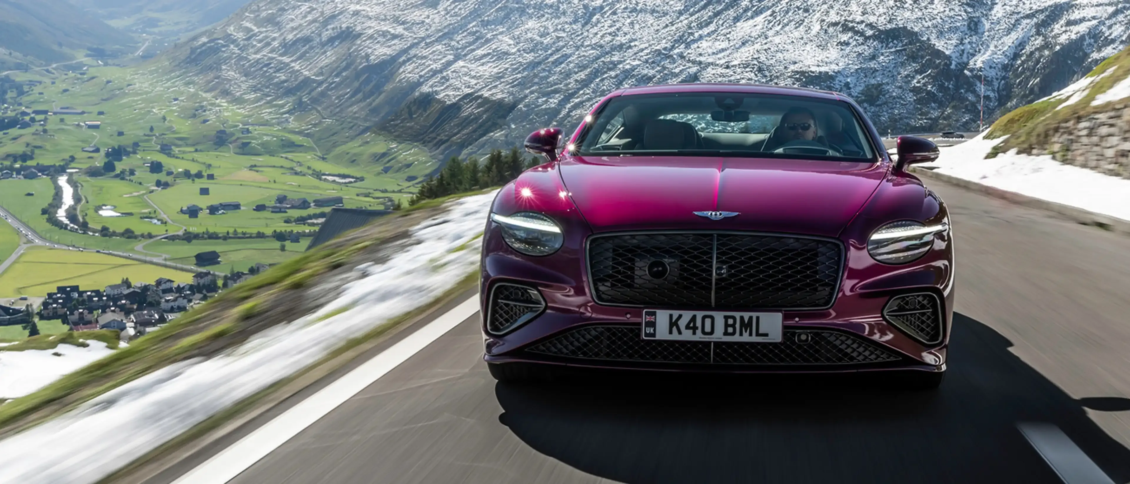 Front view of a magenta Bentley Continental GT driving a mountain pass, snow-lined road and alpine valley below.